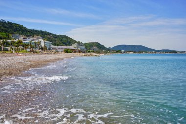 otel ve arka planda bazı küçük dağlar, mavi gökyüzü ve berrak su ile Akdeniz'de plaj