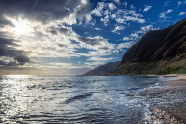 Dalgalar ile kumlu plajda bulutların arasından parlayan güneş