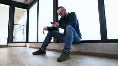 Genç adam panoramik pencere oturuyor ve ekran üzerinden cep telefonundan çevirir. Üzgün adam.