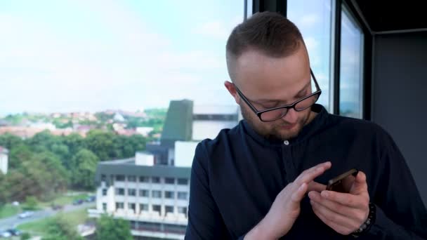 Mladý Vousatý Muž Brýlemi Zůstane Okna Vypadá Jeho Mobilní Telefon — Stock video