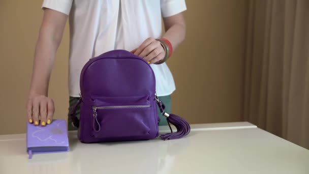 Eine junge Frau holt den Inhalt ihres lila Rucksacks hervor und legt alles auf einen weißen Tisch. Inhalt der Damentasche. — Stockvideo