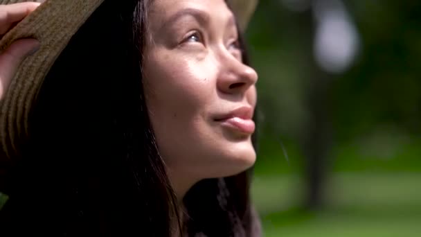 Primer plano de una chica asiática sonriente en un sombrero de verano mirando al sol y guiñando los ojos . — Vídeos de Stock