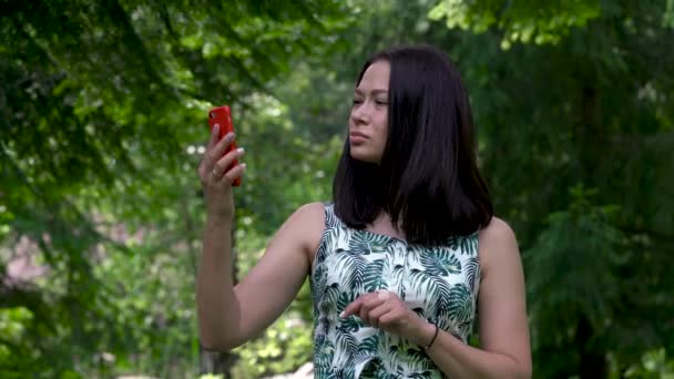 Linda chica asiática intenta conectarse a una señal de red celular en el parque. El teléfono rojo está en sus manos . — Vídeos de Stock