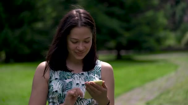 Primo piano di una donna asiatica dall'aspetto felice che cammina nel parco e digita sms sullo smartphone giallo . — Video Stock