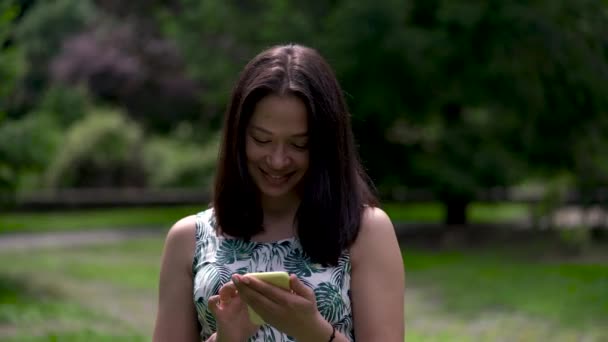 Happy looking asian woman walks in the park and types sms on the yellow smartphone. — Stock Video