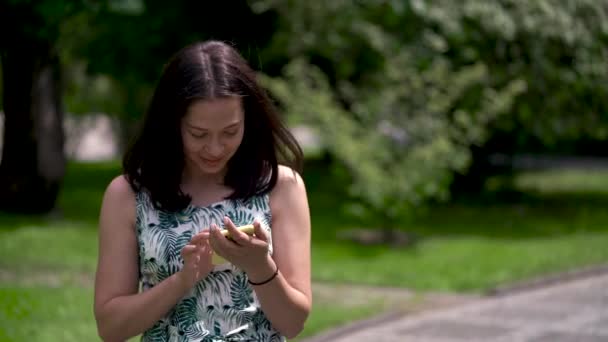 Gülümseyen Asyalı kız parkta döşeli parça boyunca gider. Onun sarı Smartphone cep telefonu ile sosyal ağlar sohbet. — Stok video