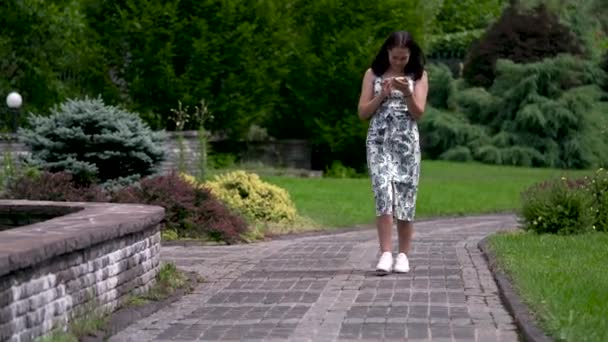 Sonriente chica asiática en un vestido de verano camina a lo largo de la pista en el parque. Charlando en las redes sociales. A toda altura . — Vídeos de Stock