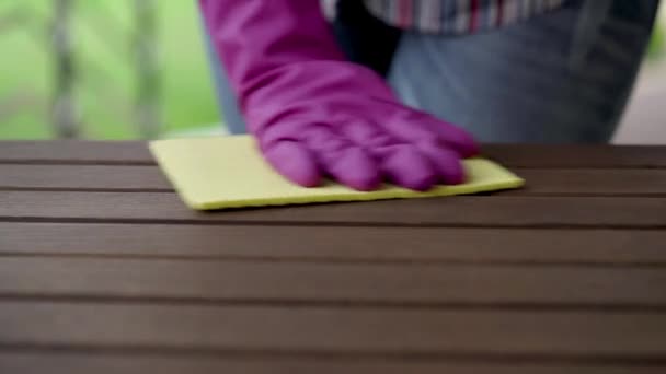 Закрытие руки в фиолетовой перчатке очищает деревянный стол на террасе желтой тряпкой. Женская фигура на заднем плане . — стоковое видео