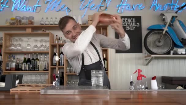 Un tânăr barman chipeș scutură un cocktail albastru într-un agitator de cocktail care stă la tejgheaua de bar. Apoi, el toarnă conținutul agitatorului într-un pahar . — Videoclip de stoc