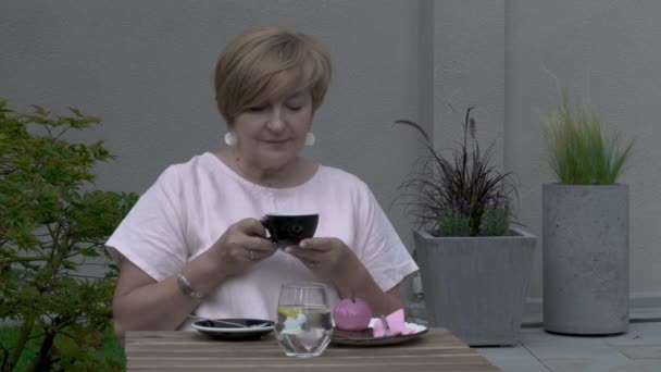 Une femme âgée tient une tasse de thé, regarde dedans et autour de la cour . — Video