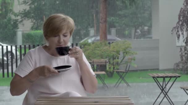 Közelről elég idős nő ízű tea, ül a teraszon heves esőzés — Stock videók