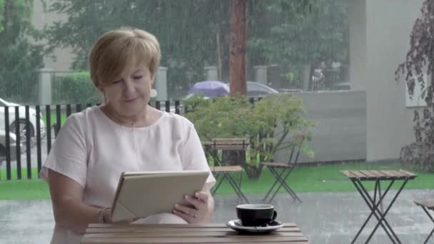 Uma mulher idosa atraente olhando para o tablet, digitando, sentado no terraço de verão em tempo chuvoso — Vídeo de Stock