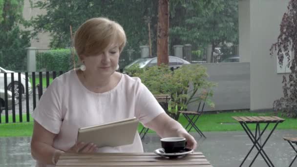 Пожилая женщина на террасе читает планшет и наслаждается чашкой чая. На заднем плане идёт сильный дождь. — стоковое видео