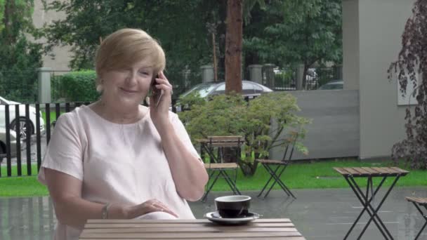 Красивая пожилая женщина счастливо разговаривает по телефону на террасе. Она улыбается и хлопает рукой. — стоковое видео