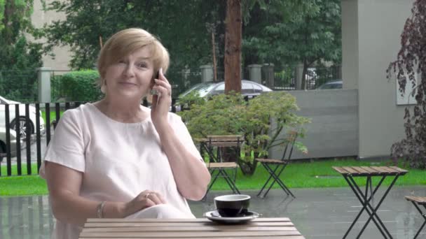 Гарна літня жінка щасливо розмовляє по телефону. Вона сидить на терасі — стокове відео