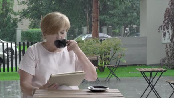 Mulher idosa no terraço lê o tablet reagindo às notícias, bebendo chá — Vídeo de Stock