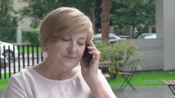 Un primer plano de una mujer muy anciana hablando por teléfono. Ella está sentada afuera en la terraza. La lluvia está cayendo . — Vídeos de Stock