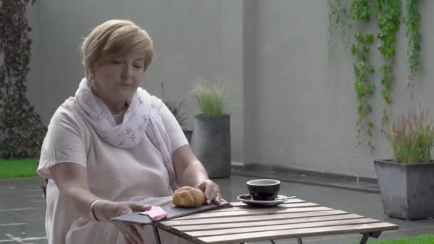 Una bella donna anziana sta mangiando un croissant e bevendo il tè. Si siede sulla terrazza. Sta piovendo. . — Video Stock