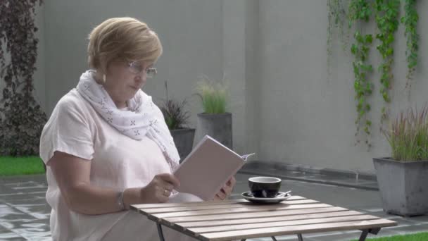 Una mujer muy anciana está sentada al aire libre en la terraza y leyendo un libro. Lleva gafas. . — Vídeos de Stock