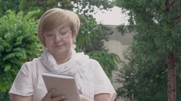 Een dolly voor een vrij oude vrouw zitten buiten en lezen van een boek. Natuur in de achtergrond. — Stockvideo