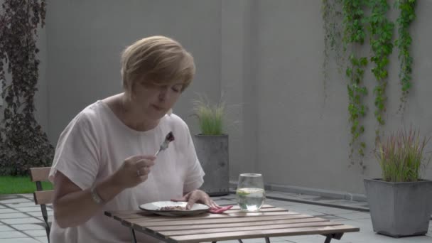 En äldre kvinna slutar äta en kaka. Hon sitter utanför på terrassen. Hon torkar läpparna och dricker lite vatten efteråt — Stockvideo