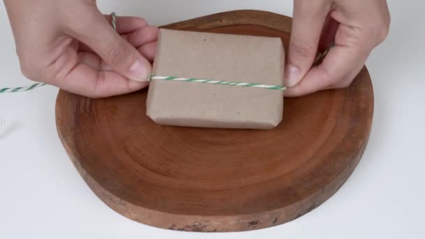 A close-up of a bar of natural soap wrapped in natural brown paper. Woman's hands fold the corners and wrap a string around the parcel, make a bow. — Stock Video