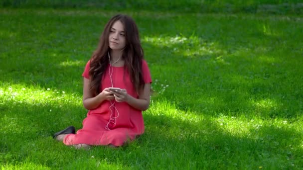 一个年轻漂亮的女人坐在草地上的特写 她戴着耳机听音乐 她在调 — 图库视频影像