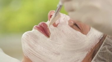 A yakın çekim kadının yüzü beyaz maskeyle kaplı. Bir fırça ile bir el daha fazla maske madde yüzünde geçerlidir.