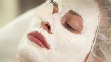 Kadının yüzüne üstten görünüm beyaz maskeyle kaplı. Bir fırça ile bir el onun burun, dudak ve çene üzerinde daha fazla maskesi uygular