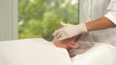 Güzellik uzmanı tarafından temizlenmiş kadının yüzüne yan görünüm. Bir el kremi bir kavanoz tutar, biraz krema alır ve alın, yanaklar geçerlidir ve cilt, boyun ve alın masaj başlar