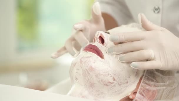 Un primer plano de la cara de la mujer cubierta de máscara blanca. Dos manos en guantes comienzan a masajear las mejillas, a lo largo de la línea de la mandíbula en círculos pequeños — Vídeos de Stock