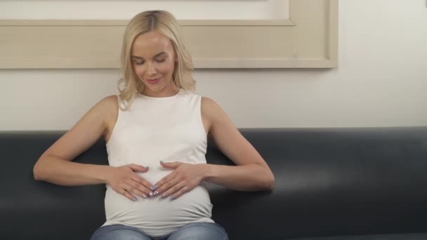Передний вид очаровательной беременной блондинки, сидящей на диване, трогающей живот и счастливо улыбающейся — стоковое видео