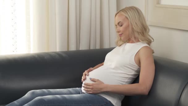 Боковой вид довольно беременной блондинки сидит на диване и гладит ее живот, она смотрит на него и улыбается — стоковое видео