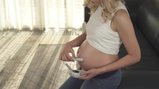 Eine Seitenansicht einer schwangeren Blondine, die den fetalen Herzschlag kontrolliert. bewegt sich die Kamera von unten nach oben. sie blickt auf den Bauch und lächelt. — Stockvideo