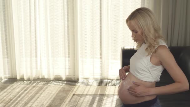 Боковой вид беременной блондинки, стоящей в комнате и втирающей сливки ей в живот. Она смотрит на живот и наносит его кругами обеими руками. — стоковое видео
