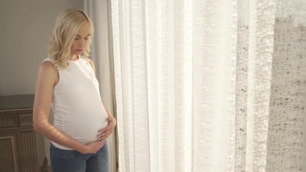 Uma vista lateral de uma linda loira em pé junto à janela e acariciando sua barriga. Ela olha para a barriga e em torno dela sorrindo feliz — Vídeo de Stock