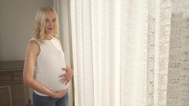 Une vue latérale d'une jolie blonde enceinte debout près de la fenêtre. Elle regarde la caméra sourire, tient une main sous le ventre et caresse son ventre avec un autre — Video