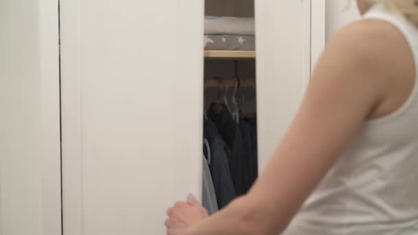 Uma foto do guarda-roupa branco fechado. Uma mulher loira aproxima-se. As mãos dela abrem as portas. Há roupas de bebé. . — Vídeo de Stock