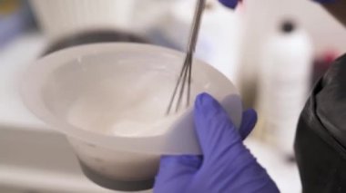 Mavi kauçuk eldiven saç boyaları ile bir renk tonu kase tutarak ellerinde yan görünüm yakından. Bir el bir çırpma teli ve renk tonu fırça ile karışımları