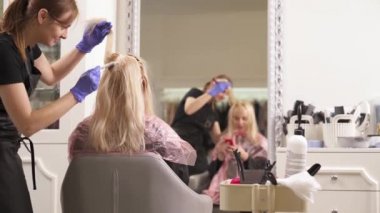 Sarışın Kuaför, sandalyede oturur. Kuaför saç boyaları ayakta soldaki kadının arkasındaki geçerlidir. Onların yansıma ayna önünde bulanık. Sarışın bir telefon tutar.