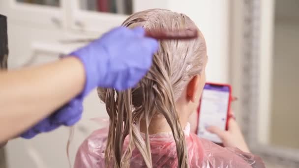 Uma mulher com cabelo tingido senta-se de volta para a câmera. O cabeleireiro pega um fio e o estica com o cabo do pente da cauda do rato. A mulher segura um telefone e digita uma mensagem . — Vídeo de Stock