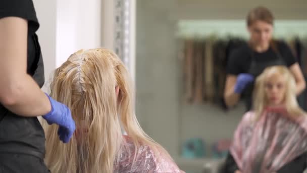Een achteraanzicht van de blonde zittend in de stoel bij de kapper met geverfd haarwortels. De kapper strekt zich uit van haar in strengen met lengte. Zij worden weerspiegeld in de spiegel voor hen. — Stockvideo