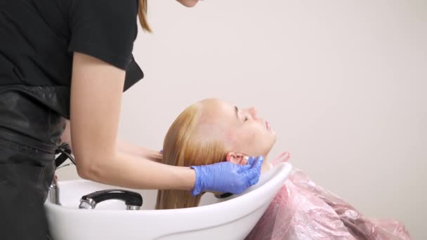 Een close-up zijaanzicht van de kapper holding een douche en wassen van de blonde haren in het bekken backwash. — Stockvideo