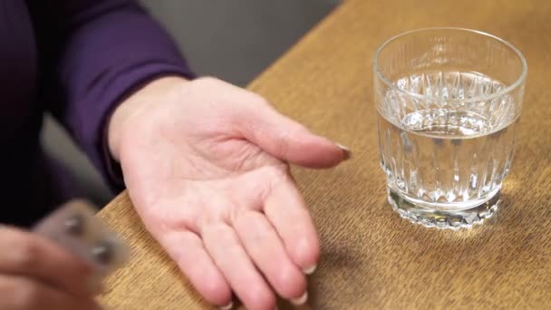 Крупный план стакана с водой, стоящей на столе. Рука старухи держит пузырь от таблеток и выпивает одну таблетку. — стоковое видео
