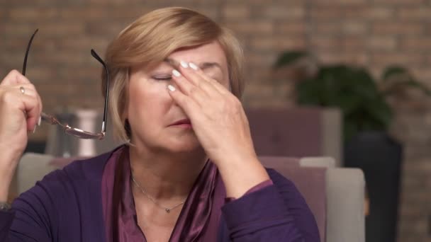 A close-up of a middle-aged woman taking off and holding glasses in her left hand. She rubs her forehead, a nose bridge, blinks eyes to rest a bit — Stock Video