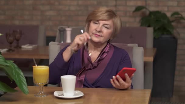 매력적인 중 년 여 자가 전화로 카페 테이블에 앉아. 그녀는 안경에 박 았 하 고 전화를 스크롤 시작 — 비디오