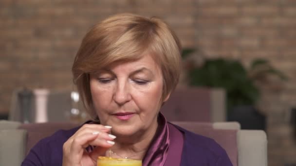 Ett porträtt av en charmig medelålders kvinna tar ett glas med apelsinjuice och dricka det genom stråna — Stockvideo