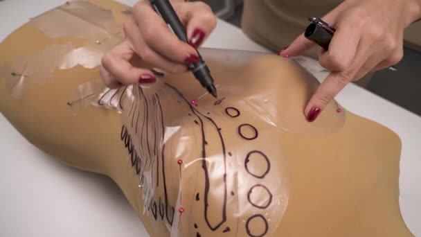 Uma visão superior das mãos do designer finalizando padrões com uma caneta de feltro no modelo de protótipo preso ao manequim deitado na mesa — Vídeo de Stock