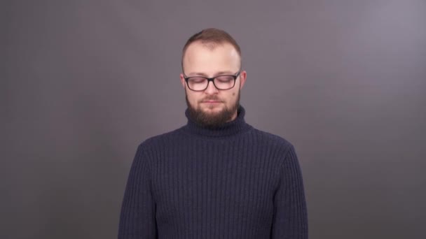 Portrait d'un jeune homme barbu à lunettes au regard coupable. Isolé sur fond gris . — Video