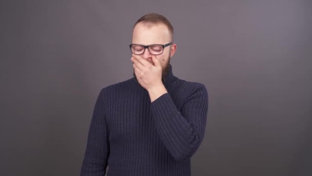 Portrait of tired yawning man with beard and glasses, dressed in a turtleneck. Isolated on grey background. — Stock Video